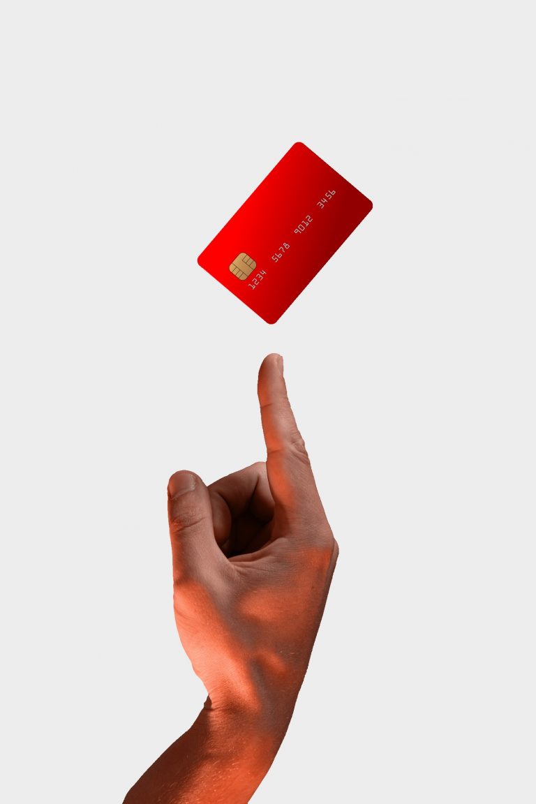 Nahaufnahme männliche Hand zeigt mit dem Zeigefinger nach oben auf schwebende rote Debitkarte auf grauem Hintergrund debit-karte.com